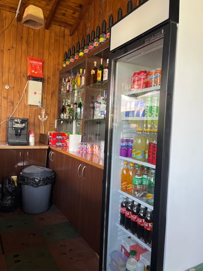The image presents a scene inside a room, possibly a bar or a shop, in Valchi Dol, Bulgaria. The primary focus is a glass-fronted refrigerator stocked with various beverages, including canned and bottled soft drinks such as Coca-Cola, Fanta, and Schweppes. The refrigerator is black-framed and situated against a white wall. Beneath the refrigerator are wooden cabinets, where additional items, including Lush brand products and disposable cups, are stored. A gray trash can with a black liner is next to the cabinets. Adjacent to the fridge is a display case filled with bottles of alcohol. On the wooden wall, a woven basket and a fire blanket, next to a medical kit, are mounted. Below is a grey espresso machine. The floor is tiled with a brown and green patterned tile.