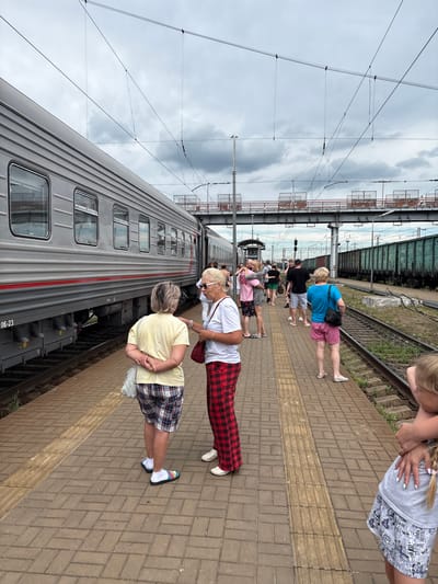The image depicts a railway platform in Povorino, Russia, on an overcast day. A passenger train is stopped at the platform, and several individuals are visible. In the foreground, two women stand facing each other, engaged in conversation. One woman, wearing a yellow t-shirt and plaid shorts, has her hands clasped behind her back. The other, in a white t-shirt and red plaid pants, holds a red bag. To the right, a woman in a blue t-shirt and pink shorts stands with her back to the camera, looking towards the railway tracks. Further down the platform, a group of people are seen, some in casual attire, suggesting they are waiting or disembarking. The background reveals more railway tracks with freight cars and an elevated pedestrian bridge or overpass. The platform itself is paved with brick, marked with tactile paving. Overhead, power lines for the electric train are suspended.
