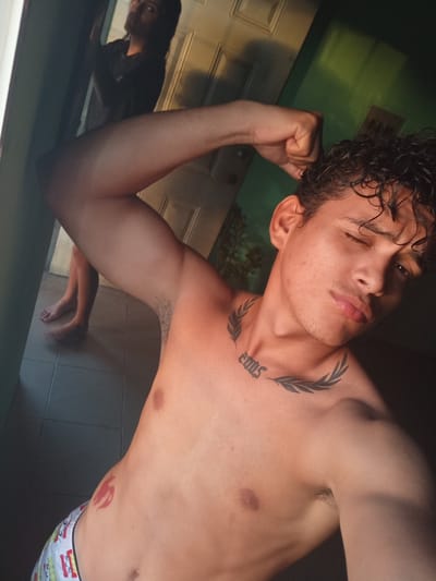 A young adult male, appearing to be in his late teens or early twenties, poses for a selfie in what appears to be an indoor environment, possibly a residential setting in La Guardia, Venezuela. The male is shirtless, revealing a tattoo on his chest that reads "2005" with a laurel wreath design above and below it, and another red tattoo on his left side resembling a stylized bird or dragon. He is wearing patterned boxer briefs.
The background shows a doorway and what appears to be a reflection in a mirror or window of another person standing. The lighting suggests it is late afternoon or early evening, with sunlight streaming in from the left, casting shadows and highlighting the contours of the male's body. The floor is tiled. The overall impression is a casual, possibly impromptu, photograph.