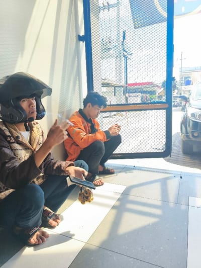 Two men sit on the floor near a metal gate outside a building in Bojongsoang, Indonesia. One man is wearing a black helmet and is holding a cell phone in his hand. He is wearing a brown jacket and dark blue jeans. The other man is wearing an orange jacket and dark pants. He is looking at the phone in his hand. A stuffed animal sits on the floor between the two men. The scene is lit by natural sunlight coming from the right. There is a car parked outside the gate. The gate has a wire mesh and is made of metal. There is a shadow of the gate and the men on the floor. The building's exterior wall is white.