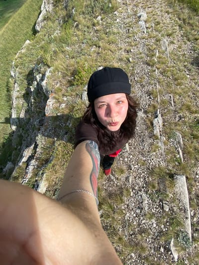This is a first-person perspective selfie video taken outdoors in a rocky, grassy area. The subject, a person with dark hair, is wearing a black beanie and a dark shirt. They have a tattoo on their arm and are looking up at the camera with a playful expression, puckering their lips. The background consists of layered rock formations and sparse green grass, suggesting a natural, possibly mountainous or hilly, environment. The lighting indicates it is daytime, with bright sunlight casting shadows. The terrain appears somewhat rugged and uneven. There is no visible text in the video. The overall mood seems casual and adventurous.