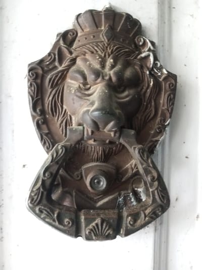 A tarnished, ornate lion head door knocker affixed to a white surface. The lion wears a crown, with intricate detailing on the mane and facial features. The knocker mechanism hangs below the lion's snout. The entire piece has a weathered, antique appearance, with signs of wear and discoloration. A peephole is present within the lower half of the door knocker.