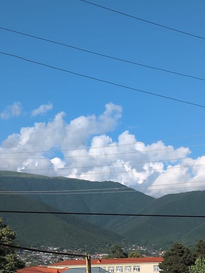 This media file shows a scenic view of a mountainous landscape near Şəki, Azerbaijan. The sky is a vibrant blue, dotted with fluffy white clouds. Several thin wires, likely part of a power line or cable system, stretch diagonally across the frame, obscuring parts of the view.

On the right side of the image, a lush green mountainside descends towards a cluster of buildings, suggesting a town or village nestled at the foot of the mountains. The architecture of the buildings appears to be of a warm, earthy tone, with visible windows. Further up the slope, the dense foliage of trees forms a rich green carpet. The presence of these human settlements against the natural grandeur of the mountains evokes a sense of harmony between civilization and nature.

The lighting suggests it is daytime, with the sun likely high in the sky, casting clear shadows and illuminating the clouds. The overall atmosphere is serene and peaceful, capturing a beautiful day in a picturesque location. No people are visible in the frame, and there is no visible text.