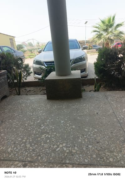 This image depicts an outdoor scene in Gwammaja, Nigeria, on what appears to be a bright, hazy day. The foreground features a light-colored terrazzo floor composed of various sized aggregate. A prominent, tall, light-grey cylindrical column rises from a textured, rectangular concrete base, centrally located in the lower mid-frame. To the left of the column base, a decorative concrete block planter is partially visible, while to its right, a small garden bed contains dark green shrubs and two upright snake plants (Sansevieria trifasciata) in exposed soil.

Beyond the foreground, two sedans are parked on a paved surface. A light gold or silver Honda Accord (likely a 9th generation model) is parked facing the viewer, positioned behind the central column. To its left and slightly further back, the rear portion of a dark grey or silver sedan is visible. In the background, a light-yellow building with a patterned screen is seen on the left. The right background features a large palm tree with green fronds, and behind it, a tall pole equipped with a solar panel and a street light fixture. Another faint red vehicle is partially visible further down the street to the right. Power lines crisscross the upper left and central sky. The distant background on the left shows an undeveloped, dusty area with sparse vegetation and what may be distant structures or hills, with a faint plume of smoke or dust in the air. No individuals are present, and no actions or interactions are depicted.