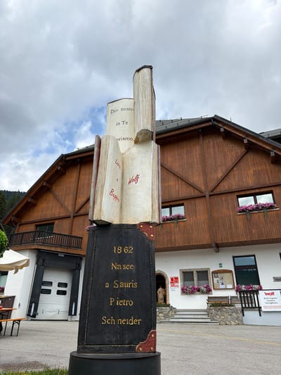 The image shows a monument in the shape of an open book, placed outdoors in front of a wooden-clad building. The monument has text inscribed on its pages and base. The text on the open pages of the book appears to be in Italian, with some words written in red. The base of the monument is black with gold lettering, which reads "1862 Nasce a Sauris Pietro Schneider". This indicates the monument is dedicated to Pietro Schneider, born in Sauris in 1862. The building behind the monument is a multi-story structure with a dark wooden facade and a gray roof. It has windows with flower boxes filled with pink flowers, and a balcony on the upper level. In the foreground, there is a paved area with some grass and yellow flowers. To the left of the monument, part of a white tent and an outdoor table with benches are visible. Further to the left, there is a large garage door. To the right of the monument, the entrance to the building is visible, with steps leading up to a door and windows adorned with flower boxes. A sign with the "Wolf" logo is also visible on the right side of the building. The sky is overcast with clouds. Sauris / Zahre is a municipality in the province of Udine, in the Friuli-Venezia Giulia region of northeastern Italy. The scene depicts a public space, likely a town square or a plaza, in Sauris.