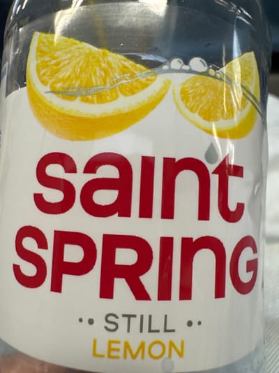 The image is a close-up of a clear plastic bottle of Saint Spring Still Lemon flavored water. The bottle label features two lemon slices and the brand name "saint SPRING" in large red letters. Below the brand name, in smaller gray text, are the words ".. STILL ..". At the bottom of the label, in yellow text, are the words "LEMON". The background of the image is blurred and appears to be a light-colored fabric surface, possibly clothing or upholstery. There are no people or any discernible city features visible. Therefore, the location in Selyatino, Russia cannot be determined from the provided image.