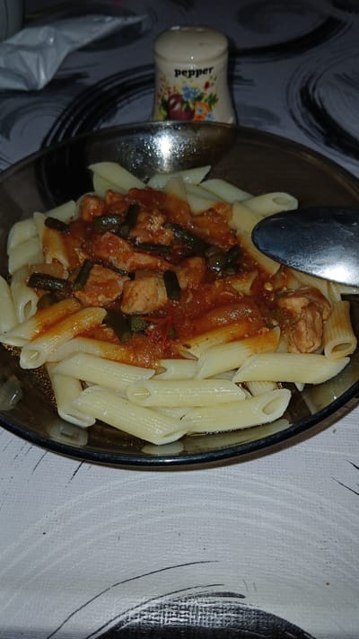 In Kolyvan, Russia, a close-up shot captures a dish of penne pasta served with a tomato-based sauce containing pieces of chicken and green beans. The pasta is arranged in a bowl, with a spoon resting on the right side of the frame. In the background, a salt shaker labeled "pepper" is visible. The scene is set on a table covered with a patterned tablecloth.