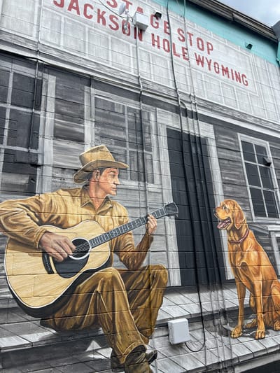 The image shows a mural on the side of a building in Jackson, Wyoming, United States of America. The mural depicts a man in a cowboy hat playing an acoustic guitar, with a golden retriever sitting next to him. The man is wearing a brown long-sleeved shirt and pants, and the dog is wearing a pink collar. Behind them, the mural is painted to resemble the facade of a building with windows and doors. The words "STAGE STOP JACKSON HOLE WYOMING" are painted in red letters at the top of the mural. The building itself is made of brick, and the mural is painted directly onto the bricks. The sky is overcast.