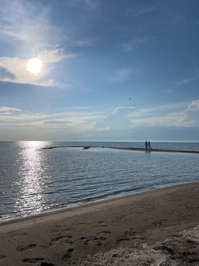 This image depicts a serene beach scene with a wide expanse of calm water meeting dark sand. The sky is a clear, bright blue with scattered wispy white clouds, suggesting a pleasant, sunny day. In the distance, two figures can be seen walking along the waterline, their forms silhouetted against the shimmering water. The sun is positioned high in the sky, casting a bright glare on the water's surface, creating a sparkling effect. The dark sand is marked with numerous footprints, indicating recent activity. The overall mood of the image is peaceful and tranquil, evoking a sense of calm and relaxation at the seaside.