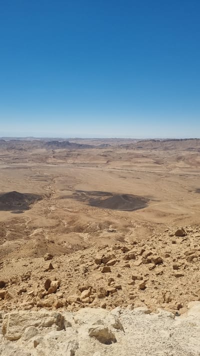 This media file shows a panoramic view of a desert landscape. The image is oriented vertically, with the sky taking up the left half and the desert terrain on the right. The sky is a clear, bright blue, transitioning to a lighter shade towards the horizon, suggesting daytime.

The desert scene is vast and arid, characterized by rolling hills, dry riverbeds, and sparse vegetation. The ground is predominantly sandy and rocky, with varying shades of brown and beige. Shadows are cast across the landscape, indicating the sun is at an angle, likely mid-morning or late afternoon. In the foreground, the terrain consists of rough, textured rock formations with visible cracks and rough surfaces. Further into the distance, the land flattens out into a wide expanse, with distant mountains or mesas forming a rugged skyline. A thin, lighter-colored line, possibly a road or a dry watercourse, can be seen snaking through the middle ground. The overall impression is one of a desolate, natural environment.

There are no people or man-made objects explicitly visible in the scene, besides the potential road. The weather appears to be clear and sunny. The location is identified as Mizpe Ramon, Israel, which is known for its desert landscapes, including the Ramon Crater. The emotion conveyed is one of vastness and natural beauty, with a sense of solitude. No text is visible in the image.