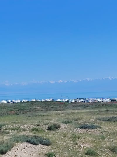The image depicts a wide landscape under a clear blue sky. In the foreground, a rocky, grassy terrain stretches across the frame. In the mid-ground, a line of white yurts, characteristic of nomadic dwellings, is visible along the shore of a body of water, likely a lake. Beyond the yurts, the water stretches out, and in the distance, a snow-capped mountain range forms a dramatic backdrop. A single boat can be seen on the water. The scene is tranquil and suggests a rural or semi-nomadic setting, possibly a tourist camp or a permanent settlement in a natural environment. The overall impression is one of vastness and natural beauty.