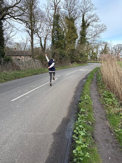 An individual is captured mid-stride, running along a rural asphalt road in North Norfolk, United Kingdom, under an overcast sky. The person, viewed from behind, wears a dark blue sleeveless vest over a light-colored long-sleeved top, grey athletic leggings, and brown running shoes. White over-ear headphones are visible. Their right arm is raised vertically, hand partially clenched, while the left arm is positioned lower and bent.

The road features a white dashed center line and the word "SLOW" painted in white ahead of the runner. To the left of the road, a light-colored wooden picket fence borders dense vegetation, consisting primarily of bare deciduous trees and some evergreens. A traditional thatched cottage with white window frames is visible through the trees in the background. To the right of the road, a narrow dirt verge with green ground cover transitions into a thick stand of tall, dry, light brown reeds. The scene suggests a late autumn or winter environment.