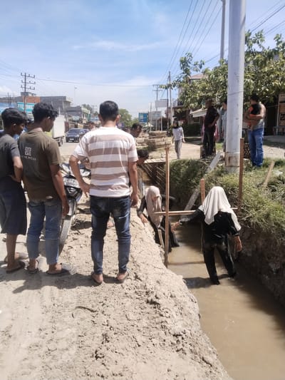 In Manetunong, Indonesia, a group of people are gathered at a construction site. Some stand on the edge of a newly dug trench filled with muddy water. Two women, wearing headscarves, are inside the trench, seemingly working on something. Other people are watching from a short distance away, including a person holding a baby. A motorcycle is parked near the edge of the trench where the standing group is located. In the background, there's a street with buildings and power lines under a partly cloudy sky.