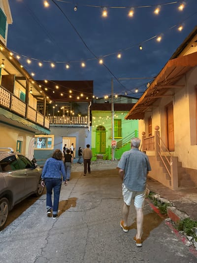 A street scene at dusk features string lights overhead. Buildings line the street, painted in various bright colors. Several people walk along the sidewalk, and a car is parked on the right side. An elderly man walks in the opposite direction of the other people. The pavement shows cracks and imperfections.