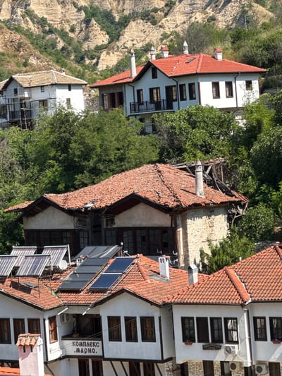 This image depicts a multi-tiered view of traditional architecture in the city of Melnik, Bulgaria, set against rugged, light-colored sandstone hills and dense green vegetation under a clear sky.

In the upper background, eroded, arid hills with visible geological strata are partially covered by sparse greenery. The midground features several houses nestled among trees. On the upper left, a white-walled house with a brown-tiled roof is partially obscured by scaffolding, indicating ongoing construction or renovation. To its right, a well-maintained white house with a vibrant red-tiled roof displays multiple decorative white chimneys, dark-framed windows, and two balconies with black railings.

Below these, a partially dilapidated house with a reddish-brown tiled roof shows significant damage on its right side, exposing wooden roof structures. It features stone and plaster walls, dark-framed windows, and a prominent grey chimney with a decorative top.

The foreground presents a cluster of interconnected white-walled houses with red-tiled roofs. Multiple solar water heaters, including both evacuated tube and flat-plate collectors, are installed on the roofs. One section displays a sign reading "КОМПЛЕКС .МАРИО." (Complex .Mario.) below which a small balcony holds a figure. An air conditioning unit is mounted on the wall. Another section features a sign reading "САМОСТОЯТЕЛНИ СТАИ" (Independent Rooms / Self-contained Rooms) and multiple dark-framed windows, with two air conditioning units visible on the lower wall. Several chimneys, including one with a traditional clay cap and others with metal caps, protrude from the roofs. The lower portions of these houses are constructed with stone.