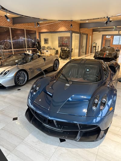 An indoor, vertically oriented image captures a luxurious car showroom in Greenwich, United States, featuring multiple Pagani supercars. The prominent subject in the foreground is a stunning dark blue Pagani Huayra, showcasing its intricate carbon fiber body panels, quad headlight design, and aggressive front splitter. Its cockpit is partially visible through the windshield, revealing the detailed interior.
Behind the dark blue Huayra, a silver or light grey Pagani Huayra is partially visible, displaying its unique wheel design and sleek profile. Further into the background, another dark-colored car, likely another Pagani, is also on display.
The setting is a high-end showroom with a clean, modern aesthetic. Features include light-colored tiled flooring, exposed brick walls, and large glass partitions separating different display areas. Overhead lighting illuminates the space, creating a polished ambiance. The cars are static, positioned for display or storage, suggesting an exhibition or private collection.
Notable details include the widespread use of visible carbon fiber on the main Pagani, highlighting its lightweight construction and exotic nature. The overall scene exudes luxury and automotive excellence. In the background, on a brick wall, a circular logo or sign is visible, potentially bearing the "PAGANI" name, though difficult to read clearly due to distance and image orientation. The scene suggests a specialized dealership or a collector's garage dedicated to hypercars.