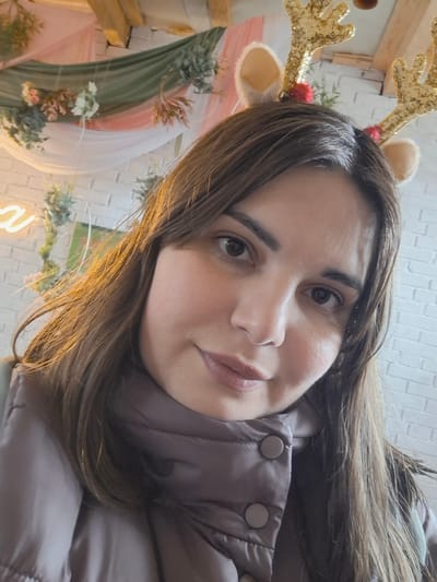A close-up portrait captures a woman taking a selfie in an indoor setting in Ludorvai, Russia. The subject, a woman likely in her 20s or 30s, has long, dark brown hair and gazes directly at the camera with a calm, pleasant expression and a subtle smile. She is dressed in a muted greyish-brown puffer jacket, featuring two visible snap buttons on the front. Adding a festive touch, she wears a headband adorned with prominent, golden glittery reindeer antlers, each decorated with a small red pom-pom.
The background suggests a decorated space, possibly for a celebration or gathering. A white brick-patterned wall forms the base of the setting, overlaid with decorative elements. These include soft, sheer drapes in pink and light green, interwoven with artificial foliage and small pink and white flowers, creating a festive garland effect. A portion of a yellow neon sign, appearing to be in cursive script, is visible in the upper background, casting a warm glow. Wooden elements, possibly shelving or part of a structure, are also visible to the right. The lighting indicates an indoor setting, likely with artificial illumination, contributing to a warm and inviting atmosphere. No discernible text is clearly visible on the neon sign.
