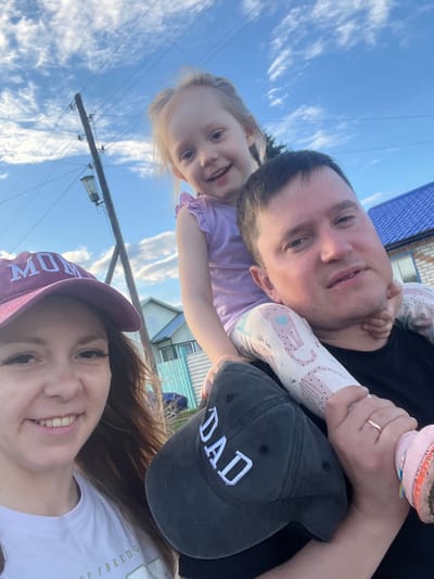 In Mal'tsevo, Russia, a close-up selfie shows a family of three in the late afternoon, with a bright blue sky and scattered clouds. The image is framed from a low angle, looking up at the subjects.
A young girl with blonde hair is perched on a man's shoulders. She wears a light purple top with ruffled sleeves and white leggings adorned with pink and blue cheetah-like prints. The man, presumably her father, has short brown hair and wears a black t-shirt. A dark baseball cap with the word "DAD" embroidered in white is placed on his head.
To the left, a woman with long brown hair is visible, wearing a purple baseball cap with "MOM" embroidered in white. She is smiling and looking towards the camera. She wears a white t-shirt with a green inscription that is partially obscured.
In the background, a wooden utility pole with a lamp attached stands to the left, with power lines extending across the sky. Behind the family, a residential area is visible with houses featuring blue and light-colored roofs, and a light blue fence. The overall scene suggests a casual outdoor family outing.