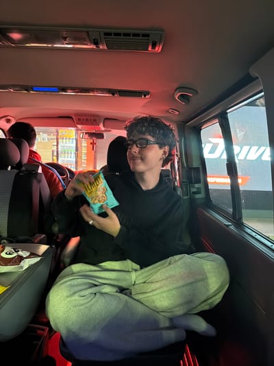 A person with short curly hair and glasses is seen inside a vehicle, holding a bag of pretzels. They are wearing a black sweatshirt and gray sweatpants, with their feet on the back seat. In the foreground, there is a headrest and a bag of chips. A person with dark hair sits in front of the person eating pretzels. In the background, a window reveals a rainy scene with the sign "DRIVE TRANSFORMA." Based on the sign, this could be at or near a gas station or rest stop with car services, although the exact location within Stara Pazova Municipality, Serbia, is not clear from the visual information alone. The inside of the vehicle has a sunroof and air conditioning vents.