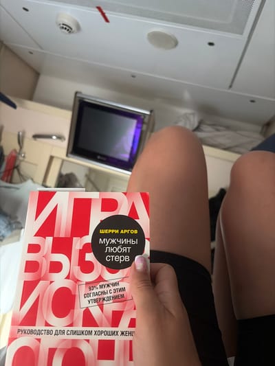 The image shows a close-up of a book titled "Мужчины любят стерв" (Men Love Bitches) by Sherry Argov. The book is red and white with large, bold lettering. A person's hand with a manicured nail is holding the book. The background appears to be an interior, possibly a train compartment or a small room, with a white ceiling, a dark screen, and some bedding. There is no indication of the city of Syzran, Russia.