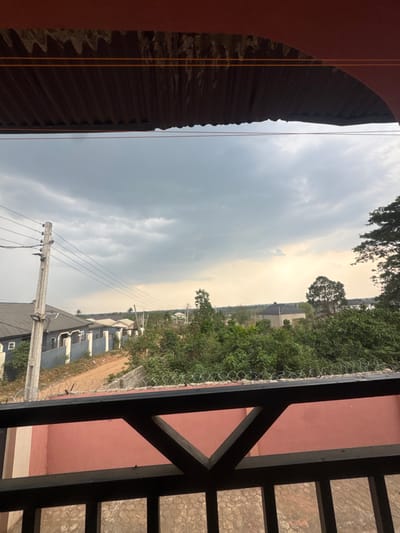 The image depicts a panoramic view of a residential area in Gbongan, Nigeria, captured from an elevated position, likely a balcony. The foreground features the underside of a dark, corrugated metal roof above, with two parallel orange wires stretched horizontally beneath it. Below the main visual field, a dark metal balcony railing with a repeating triangular and rectangular pattern is visible. Immediately behind the railing, a segment of a pinkish-red wall is present, topped with a line of barbed or razor wire.
The midground reveals a sloped dirt road on the left, flanked by several single-story residential buildings with grey roofs and light-colored walls, some secured by perimeter fences. A prominent light-colored utility pole with multiple overhead power lines stands vertically on the left. The central and right midground are dominated by dense, green vegetation, including numerous trees and bushes.
The background extends to a distant horizon line, where more scattered structures are visible amidst further vegetation. The sky is largely overcast, filled with varying shades of grey clouds, and exhibits a lighter, hazy band near the horizon on the right, suggesting a low sun or specific atmospheric conditions. No individuals or discernible actions are present in the scene.