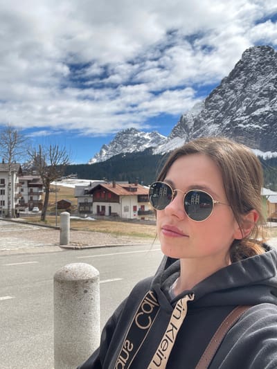 A photograph captures a person wearing a black Calvin Klein hoodie and sunglasses, with a mountainous backdrop and houses characteristic of Sappada, Italy. The mountains are snow-capped. The sky is blue with puffy clouds. Houses are visible in the reflection of the sunglasses. A cylindrical concrete bollard is partially visible in the foreground.