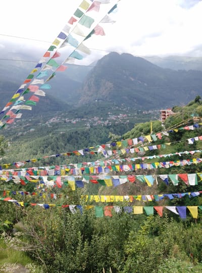 The image depicts a scenic mountain landscape in Baragran, India, adorned with numerous strings of colorful prayer flags. The flags, in shades of blue, yellow, red, green, and white, are strung across the foreground, fluttering in the breeze. They are set against a backdrop of lush green hills and a hazy sky, suggesting a mountainous region. In the mid-ground, a village or town is visible nestled on the slopes of the mountains, with buildings appearing in various colors. The time of day appears to be daytime, with natural light illuminating the scene, though the sky's brightness suggests it could be late morning or early afternoon. The weather seems mild, indicated by the clear visibility of the landscape and the gentle movement of the flags. The overall impression is one of tranquility and spiritual significance, often associated with prayer flags. There is no visible text in the image.