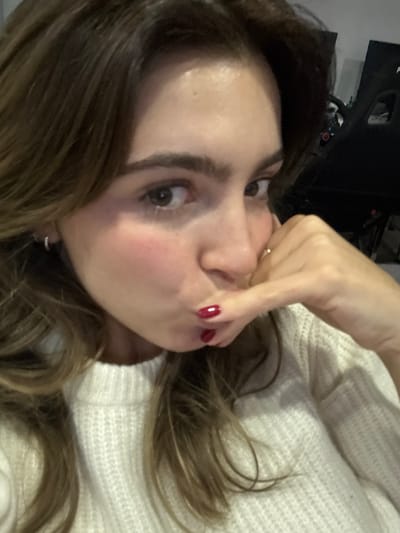 The image captures a close-up, slightly tilted view of a young person, likely female, in what appears to be an indoor setting. The individual has medium-brown, long, wavy hair that is spread out around their head. They are wearing a light-colored, possibly white or cream, knitted sweater.
The person's face is prominent, featuring wide, dark eyes looking directly at the viewer. One hand, adorned with red nail polish on the fingers and a simple ring on one finger, is pressed against their face, playfully squeezing the cheeks or mouth, creating a slightly exaggerated expression. There's a subtle blush visible on the cheeks. The background is dark and indistinct, suggesting an interior space, with some blurry shapes that could be furniture or other objects. The overall mood conveyed is playful and casual. There is no visible text in the image.