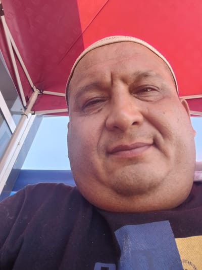 An extreme close-up selfie captures a man from the chest up, wearing a dark crewneck sweatshirt. He has a light-colored skullcap on his head and his face is turned towards the camera. The background shows a vibrant red canopy overhead, supported by white poles, suggesting an outdoor setting, possibly under a tent or awning. To the left, a section of a building with large windows and a blue structure below is visible, implying an urban or commercial environment. The sky is a pale blue. The image was taken in the city of Gulistan, Uzbekistan.
