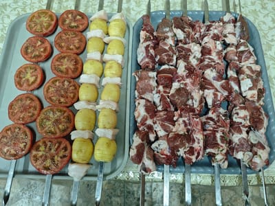 The image displays two grey trays filled with skewers of raw food, arranged on a surface covered with a light-colored, patterned tablecloth. The tray on the left contains skewers with sliced tomatoes, small, layered yellow potatoes, and cubes of white meat, likely chicken or fish. The tomatoes appear seasoned with salt and pepper. The tray on the right is filled with skewers holding cubes of raw, marbled red meat, possibly lamb or beef, interspersed with pieces of white fat and herbs. Metal skewers are inserted through the food items in both trays. The background is blurred, but the overall impression is of food prepared for grilling or cooking. The context of Goryachevodsky, Russia, suggests this might be a preparation for a traditional meal or barbecue in that region.