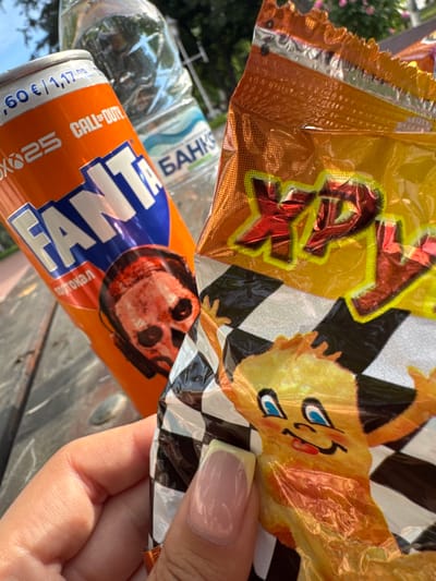 The image shows a hand holding a crinkled Fanta soda can featuring a "Call of Duty" design alongside a metallic, partially opened bag of chips with cartoon character imagery in an outdoor setting. In the background, a water bottle labeled "SAHKS" is visible. The specific location, Glojene, Bulgaria, cannot be confirmed from the visual data alone.