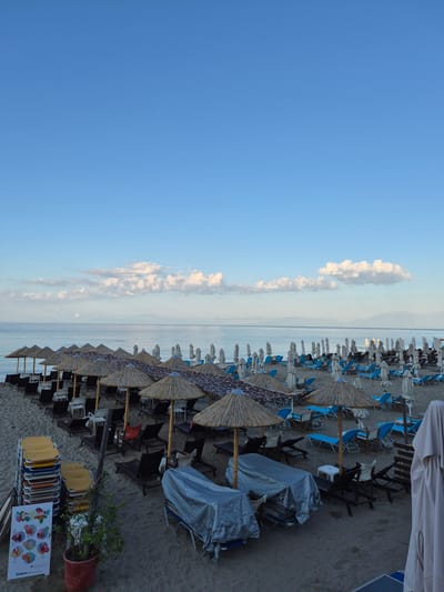 This is a high-angle, wide shot of a beach in Nea Kallikratia, Greece, likely during the early morning or late evening. The beach is mostly deserted, with rows of empty lounge chairs and umbrellas set up. The umbrellas are a mix of thatched straw and white canvas. Some lounge chairs are covered with tarps. In the foreground, there is a stack of colorful plastic chairs and a small potted plant. A sign advertising smoothies is visible to the left. The sea is calm and extends to the horizon, where hazy mountains are faintly visible. The sky is a clear blue with a few scattered clouds.