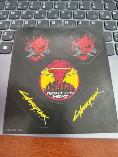 The image shows a sheet of stickers on a laptop keyboard. The stickers are black with various designs. One sticker features a red and grey demon-like mask with flames. Another is a circular sticker with a red explosion, a cityscape, and the text "NIGHT CITY HEAT" in white letters. A third sticker is yellow text that reads "CYBERPUNK 2077". There is also a small code "CPSE_ST_01_ALL" visible on the black sheet. The laptop keyboard is dark grey and appears to be a standard QWERTY layout with some Cyrillic characters visible. The setting is likely indoors, given the presence of a laptop and the lighting. No people are visible in the image.