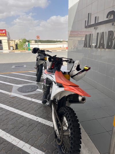 A motorcycle is parked on a paved surface with white diagonal stripes. The bike has an orange and white seat and a black exhaust pipe. A white helmet with red and yellow accents rests on the seat. The bike is positioned near a building with the name "AL JABAR" on it. A parking sign is visible in the background along with a blue sky.