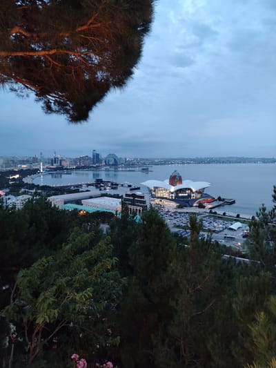 The image displays a panoramic view of the Baku skyline, specifically Sabail Raion, Azerbaijan, at dusk. In the foreground, lush green trees with pine needles partially obscure the view. The middle ground features the striking Heydar Aliyev Center, an architectural marvel with a distinctive white, petal-like roof illuminated by interior lights. Adjacent to the center is a large, busy parking lot filled with numerous cars. Further back, the Caspian Sea stretches across the horizon, reflecting the dim twilight. Beyond the sea, the cityscape of Baku is visible, characterized by a collection of modern skyscrapers, including the Flame Towers and the Baku Eye Ferris wheel. The sky is a muted blue, with scattered clouds indicating the transition from day to night. Boats are present on the water, and streetlights are beginning to illuminate the city. The overall scene conveys a sense of urban development and natural beauty juxtaposed.