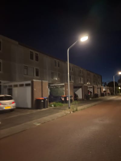 The image shows a nighttime street scene in Lelystad, Netherlands. In the foreground, a brick-paved road runs diagonally from the bottom right to the top left. To the left of the road, there is a patch of grass and a sidewalk. Further left, a white car is parked. Behind the car and sidewalk, a row of attached houses or apartments is visible. These buildings have multiple windows, and some appear to be illuminated from within. Several dark-colored trash bins with orange lids are placed along the sidewalk in front of the buildings. Two streetlights illuminate the scene, casting a bright light on the road and surrounding area. The sky above is dark, indicating it is nighttime. The overall impression is of a residential street at night.