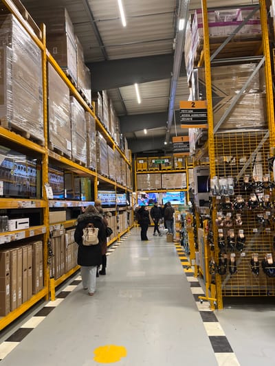 The photo captures a warehouse retail space with shelves stocked high with boxed televisions and various electronics. People stroll through the aisle, examining the displays. Yellow metal shelving units frame the scene, and overhead signs indicate product categories. Fluorescent lighting illuminates the warehouse interior, reflecting off the polished floor. A yellow marking is present on the floor.