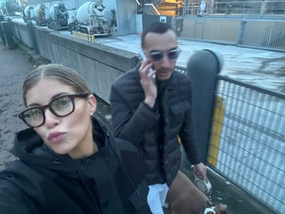 Two individuals pose in an off-kilter selfie. The woman, wearing dark-rimmed glasses and a black jacket, is positioned on the right. To her left stands a man in sunglasses and a puffer jacket, holding a dog leash attached to a light brown dog. They appear to be on a walkway featuring metal grates and yellow safety lines. Several white machines are visible in the background.