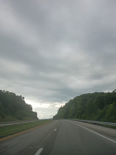 The image shows a view from a vehicle traveling on a highway in a rural area, likely near Stanton, United States of America. The road is asphalt, with visible lane markings and a yellow line on the right side. Guardrails are present on the right side of the road. Dense green trees line both sides of the highway, forming hills. The sky is overcast with thick, gray clouds. There are no people or animals visible in the image. The scene suggests a cloudy day during daylight hours, with the highway winding through a wooded landscape.