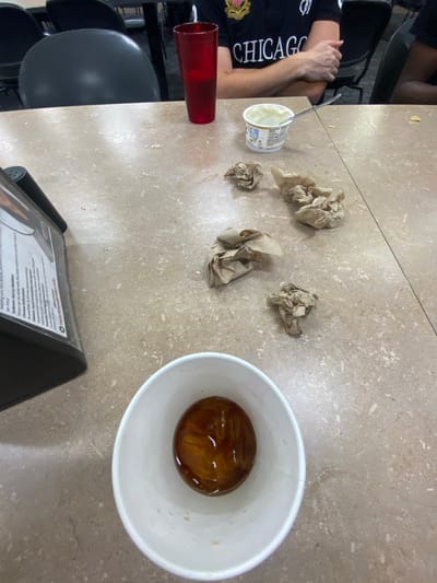 The image displays a cluttered dining table surface in what appears to be a cafeteria or dining hall environment. The precise location within Lawrence Township, United States, cannot be determined from the visual information provided.
A person, partially visible in the upper background, is seated at the table wearing a dark polo shirt with a crest-like logo and the word "CHICAGO" printed in white. Their forearms are crossed on the table. To their left, a red plastic cup holds a clear liquid, approximately half full. To their right, an open white plastic container of yogurt contains a silver spoon.
Four crumpled pieces of brown paper, consistent with used napkins, are scattered across the mid-section of the speckled, light brown tabletop. In the lower left foreground, a black plastic menu stand with white text, partially obscured, is visible, displaying words such as "Nutrition." In the center foreground, a white paper cup contains an amber-colored liquid with several ice cubes floating within it. Dark grey chairs are visible in the blurred background.