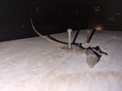 This is a close-up, nighttime photograph taken at an unspecified location in Miraverde, Spain. The foreground is dominated by a rough, light-colored surface, possibly concrete or stone, which is out of focus. Two nails are embedded in this surface. One nail, in the center of the frame, is in sharp focus and appears to be a standard metal nail with a ribbed head. The second nail, located to the right and slightly behind the first, is out of focus and appears to be rusty or made of a darker metal.

A dry, brittle, organic material, possibly a dead leaf or thin twig, is attached to the first nail and extends towards the left. This organic material is also out of focus.

The background is dark and blurry, indicating a nighttime scene. Distant streetlights and some faint lights from buildings or vehicles are visible. A road sign with some writing that is illegible due to the distance and poor lighting is present in the upper left quadrant of the image. The overall impression is that of an outdoor, urban or semi-urban environment at night, with a focus on the details of the nails and the organic material in the foreground. There are no people visible in the image.