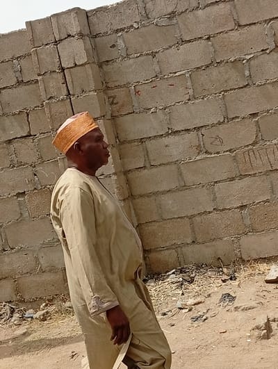 An adult male is captured outdoors in Kofar Mazugal, Nigeria. He is depicted in profile, facing to the right, wearing a light beige or khaki-colored long tunic with light-colored embroidery visible on the left sleeve cuff. A patterned orange and brown cap covers his head. The man appears to be in the process of walking or standing on an unpaved dirt surface. The ground in the lower right foreground contains scattered debris, including dry plant matter and black plastic fragments. Behind the man stands a tall, light-colored concrete block wall, constructed with unrendered, roughly mortared blocks. Faint markings, including what appears to be a red letter "Z", are visible on the wall. The upper left portion of the image shows a hazy, light-colored sky. The scene is illuminated by bright, natural light.