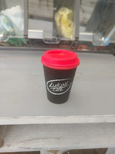 A black disposable coffee cup with a red lid sits on a light-colored surface. White text on the cup reads "Lukoil Cafe". The cup is positioned slightly to the right of the center of the frame. In the background, out of focus, are shelves with various items, including a yellow object in a plastic bag and what appear to be other cups or containers. The environment suggests an indoor setting, possibly a kiosk or a shop. The exact location within Тулаўскі сельскі Савет, Belarus is not discernible from the image itself.