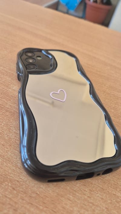 The image shows the back of a black, wavy-edged phone case with a mirrored surface. A small, light pink heart sticker is adhered to the mirrored surface, slightly off-center. The phone case is resting on a light brown, wooden surface, likely a desk or table. The background is blurred, showing hints of other objects, possibly a plant in a terra cotta pot and some blue and white items, out of focus. There are no people visible in the image. The location of Satovcha, Bulgaria is not depicted in this image.