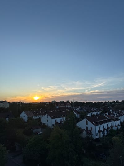 This is a medium-angle, full-frame, eye-level, outdoor, vertical shot of a residential neighborhood in Sindelfingen, Germany, during sunset. The sky is a gradient of pale blue to orange, with wispy clouds visible. The sun is low on the horizon, casting a warm glow. In the foreground, a row of white houses with red-tiled roofs lines the street. Trees and greenery are interspersed throughout the neighborhood, with some taller trees and power lines visible in the distance. The overall impression is one of a peaceful, suburban evening.