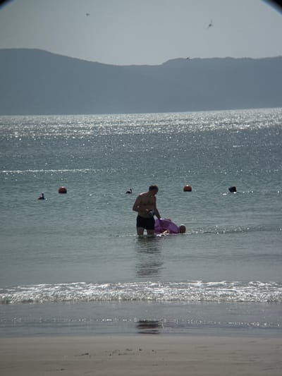 The image depicts a serene beach scene in Dong Ninh Hoa, Vietnam. In the foreground, gentle waves lap onto a sandy shore. The middle ground is occupied by the clear, calm sea, with several buoys dotting the water. In the distance, a hazy mountain range forms a picturesque backdrop.
The central focus is a man standing in the water, holding a purple inflatable. He appears to be assisting another person, possibly a child, who is partially submerged and holding onto the inflatable. The man is shirtless, wearing dark swim trunks. The water is shallow around them.
Further out in the sea, another person with a snorkel mask is visible. The sun glints off the water, creating a shimmering effect. Two birds are captured in flight against the pale sky, adding a touch of dynamic movement to the tranquil scene. The overall atmosphere is one of leisure and recreation on a beautiful day at the beach.