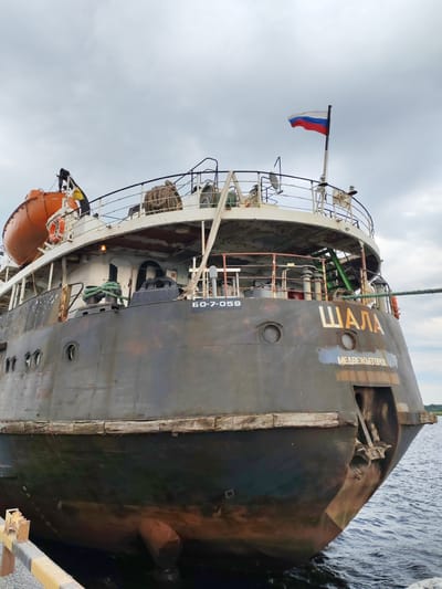 A Russian flag flies from a pole atop a weathered, dark-hulled ship docked at a pier. The ship has an orange life raft mounted on its port side. Text on the stern reads "ШАЛА" in large, light-colored letters and "МЕДВЕЖЬЕГОРСК" in smaller letters below. A registration number "БО-7-059" is also visible on the stern. The ship is partially submerged in dark water, and its hull shows signs of age and wear, with patches of rust and discoloration. A green rope is attached to a cleat on the deck and trails off to the right. The sky is overcast, with gray clouds dominating the upper portion of the image. A yellow and gray barrier is visible along the edge of the pier in the foreground. The scene appears to be in Shalsky, Russia, given the presence of Russian maritime markings and the likely context of the ship's name and homeport.