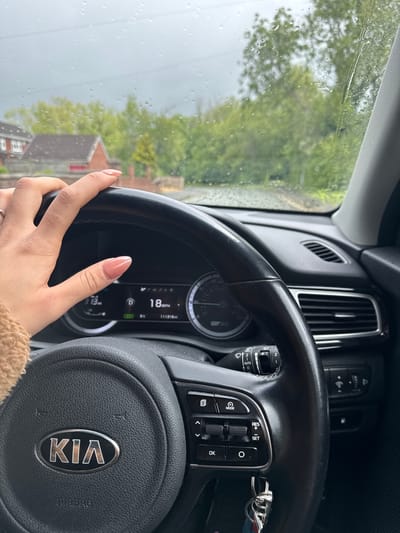 A photograph taken from inside a Kia car shows the driver's perspective while traveling through a residential area, likely in Warrington, United Kingdom, given the architecture visible through the rain-speckled windshield. The focus is on the steering wheel, prominently displaying the Kia logo. The driver's hand, adorned with light pink nail polish, is visible on the left side of the wheel. The car's dashboard is partially visible, showing a digital display reading "18 MPH" and a mileage of "11318". Rain obscures the view of the outside environment, with blurred trees and houses visible. The overall lighting suggests an overcast day.