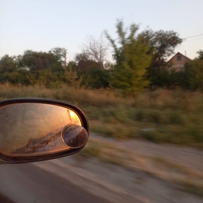 The image shows a close-up view of a car's side mirror, reflecting the setting sun and a blurred rural landscape. The mirror is dirty, with visible smudges and water spots. The text "OBJECTS IN MIRROR ARE CLOSER THAN THEY APPEAR" is inscribed on the mirror. In the reflection, a house with a peaked roof and a chimney is visible amidst trees, suggesting a rural or semi-rural setting. The background of the image is motion-blurred, indicating the car is in transit, likely on a dirt or gravel road given the texture of the foreground. The sky is a pale blue, transitioning to a warm orange hue from the setting sun. The overall impression is one of a journey through a rural area at dusk. The specific location in Sonyachne, Ukraine cannot be determined from the visual information alone.