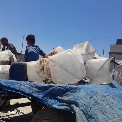 In Deir el-Balah, Palestinian Territories, a child in a blue shirt sits atop a collection of large, white plastic jerrycans filled with what appears to be sand or dirt. Beside him, a man in a dark grey shirt is partially visible, leaning forward. The jerrycans are stacked on a flatbed trailer covered with a blue tarp. The trailer's wheels and chassis are visible beneath the tarp. In the background, a clear blue sky stretches above.